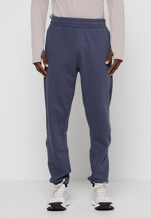 Personne portant un pantalon de jogging bleu marine, un haut à manches longues beige clair et des baskets blanches, debout devant un fond blanc uni.