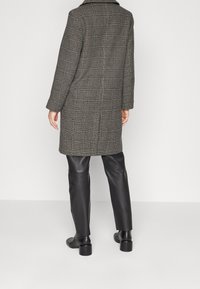 Persona che indossa un lungo cappotto a quadri, pantaloni in pelle neri e stivali neri con tacco, in piedi contro uno sfondo leggero e uniforme, vista di spalle.