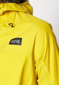 Keltainen vedenkestävä takki, jossa on korkea kaulus, säädettävä huppu ja musta "GORE"-logo rinnassa. Sileä, mattapintainen rakenne.