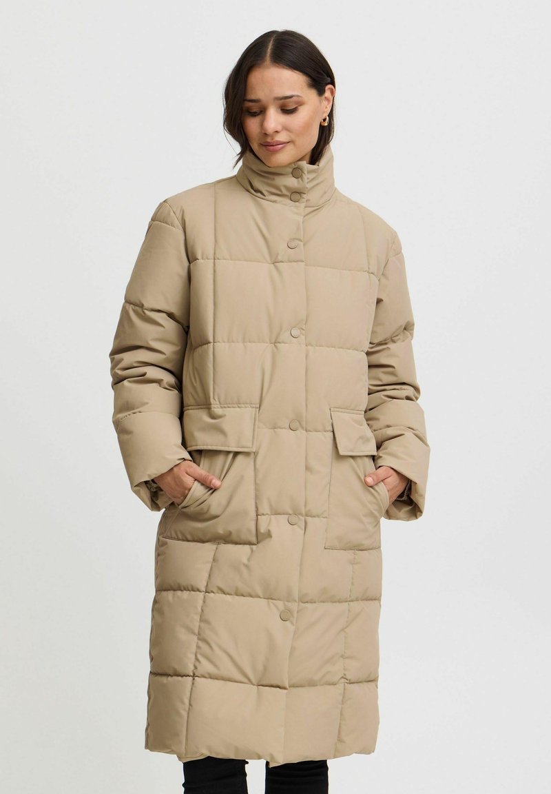 Abrigo acolchado beige hasta la rodilla con cuello alto, cierre de botones y bolsillos laterales. Presenta un diseño acolchado y una textura suave.