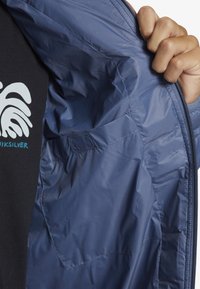 Blaue Nylonjacke mit glatter Textur, ausgestattet mit einem Reißverschluss und einer Seitentasche, teilweise geöffnet, um das Innenfutter zu zeigen.