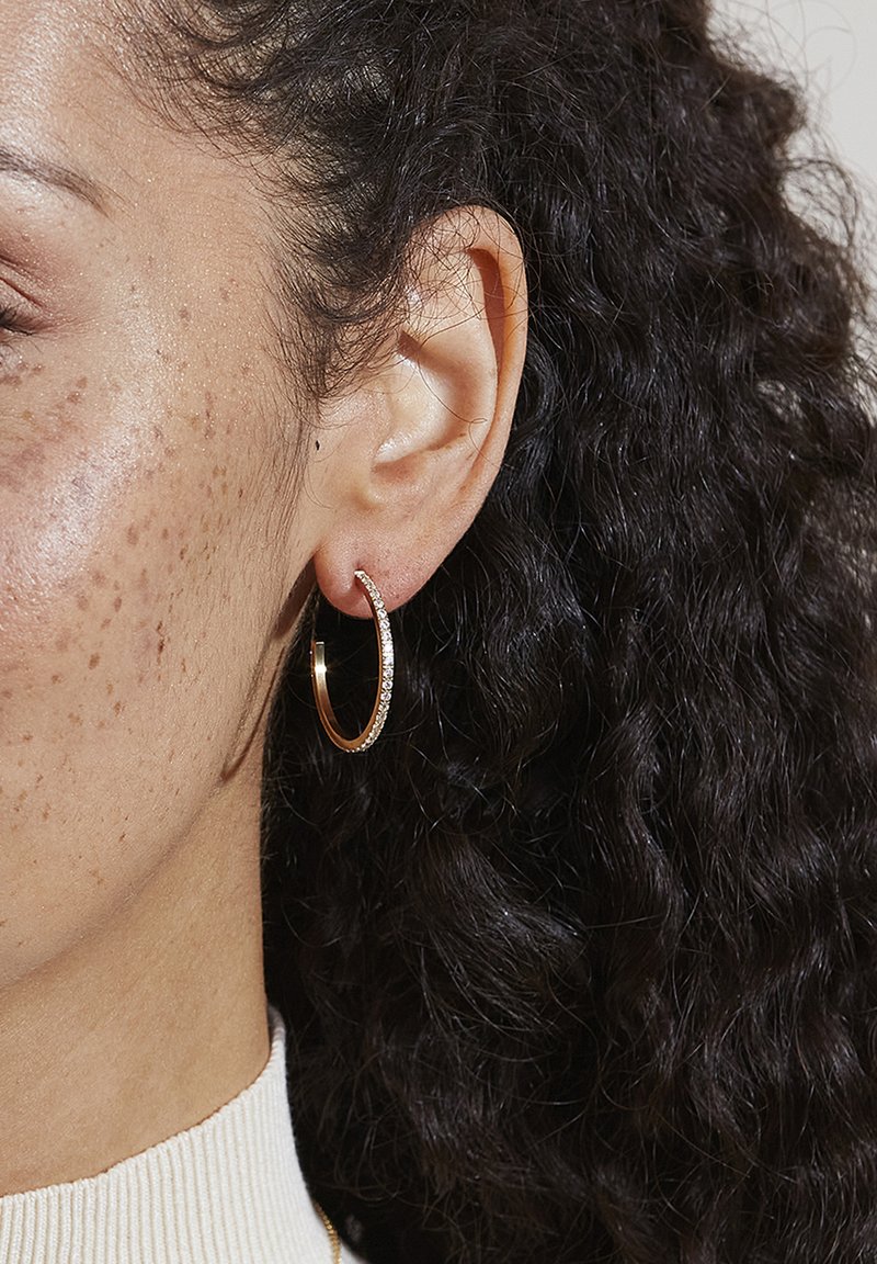 Boucle d'oreille en or avec des accents brillants, dotée d'une finition polie. Présentée sur une oreille avec des cheveux bouclés à proximité, mettant en valeur les détails.