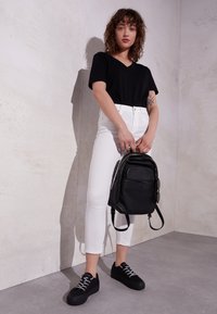 Schwarzer Leder-Rucksack mit vorderer Tasche, getragen von einer Person in einem schwarzen T-Shirt und weißen, engen Jeans, die auf einem grauen Boden steht.