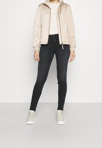 Cazadora bomber beige claro con cierre de cremallera y puños de canalé, combinada con jeans skinny negros y zapatillas gris claro.