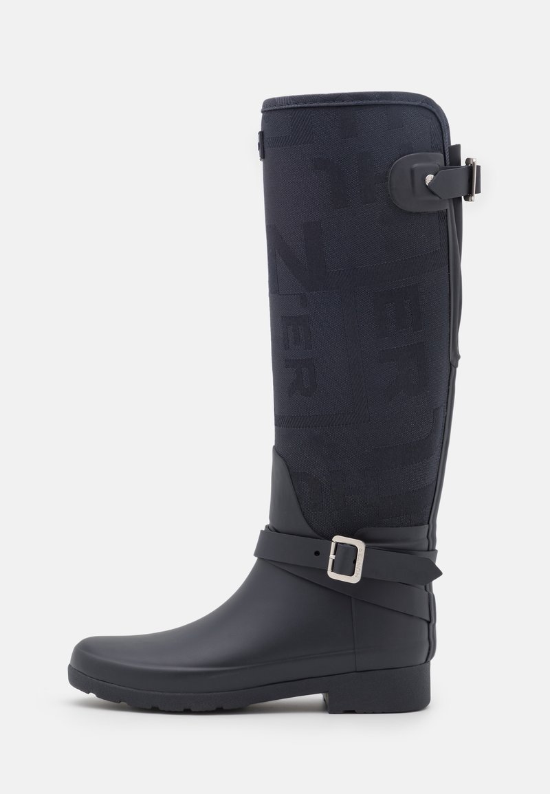 Wysoki czarny gumowy kalosz z tkaninową cholewką z delikatnie wytłoczonymi literami, paskiem na kostkę z srebrną klamrą oraz detalem klamry na górnej stronie.