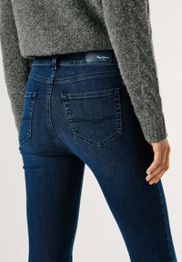 Vaqueros ajustados hechos de denim azul oscuro con una textura suave, que cuentan con bolsillos traseros tradicionales y un parche de cuero con marca en la cintura.