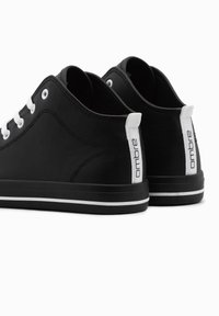 Baskets en synthétique noir avec des lacets blancs, des languettes au talon et un logo à l'arrière. Elles présentent une semelle en caoutchouc rayée blanche.