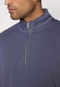 Navy zip-up sweatshirt with a high collar, featuring a silver zipper. Made from soft fabric with a smooth texture and minimal detailing.
