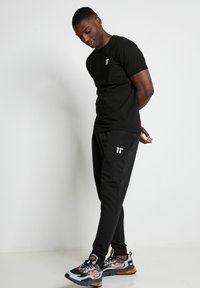 Camiseta negra con logo en el pecho, combinada con pantalones deportivos negros con marcas blancas. Zapatillas deportivas con acentos de color completan el atuendo.