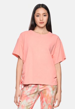 Frau mit langen dunklen Haaren, die ein lockeres, rosa kurzärmeliges Oberteil und mehrfarbige pastellgemusterte Hosen trägt, stehend vor einem schlichten Hintergrund.