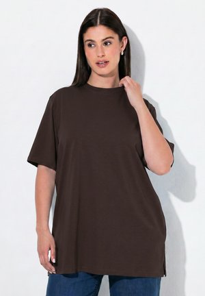 Frau mit langen dunklen Haaren, die ein lockeres dunkelbraunes T-Shirt und blaue Jeans trägt, posiert mit einer Hand nahe am Hals und schaut zur Seite.