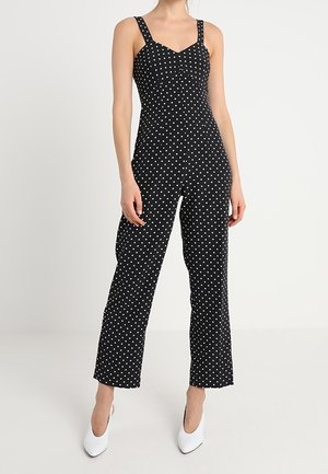 Femme portant une combinaison sans manches noire à pois blancs et des escarpins blancs à bout pointu, debout devant un fond blanc uni.