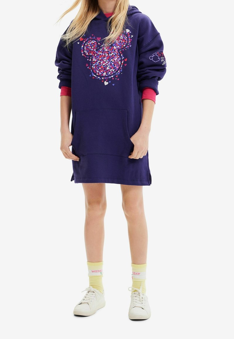 Enfant portant une robe sweat à capuche bleu marine avec un imprimé de oreilles de souris en forme de cœur colorées, des chaussettes jaunes et des baskets blanches, debout devant un fond blanc.