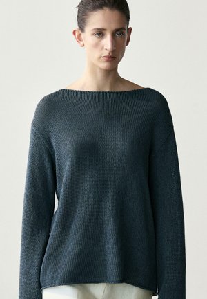 Femme aux cheveux foncés attachés, portant un pull en maille gris foncé ample et un pantalon clair, sur un fond uni.