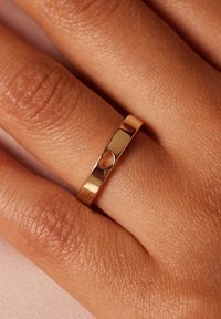 Gold ring with a polished finish, featuring a small heart cutout in the band, resting on textured skin. Simple, sleek design.