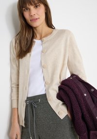 Femme portant un cardigan beige boutonné sur une chemise blanche et un pantalon gris à cordon, tenant une veste pliée de couleur violet foncé texturée.