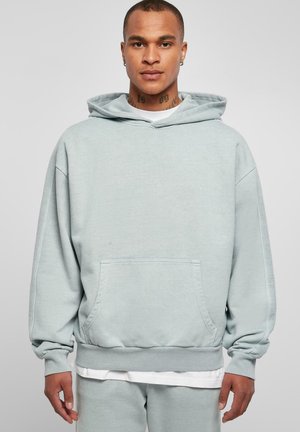 Sudadera de gran tamaño en gris claro, hecha de una mezcla de algodón, con un bolsillo frontal, puños acanalados y capucha con cordón. Lleva puesta una camisa blanca debajo.