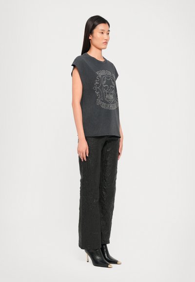 Zwarte oversized t-shirt met een frontaal schedelontwerp, gecombineerd met zwarte wijdlopende broek met textuur en zwarte enkellaarsjes met hak en gouden accenten.