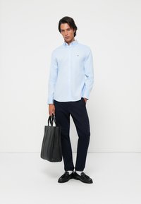 Lichtblauw overhemd met knopen, marineblauwe broek en zwart leren schoudertas. Jongenshaarstijl, zwarte schoenen en witte sokken maken de outfit compleet.