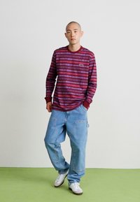 Burgundi és lila csíkos, hosszú ujjú ing, laza kék farmernadrággal párosítva. Fehér sportcipő teszi teljessé az öltözetet. Egyszerű háttér.