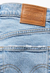 Ljusblå denimtyg med synliga sömmar; fäst brun läderlappar med präglad logotyp och detaljer vid bakre midjan.