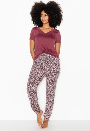 Vrouw met krullend haar staat blootsvoets, draagt een bordeaux kort mouw topje en beige luipaardprint broek tegen een witte achtergrond.