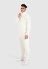 Cremefarbener Trainingsanzug aus weichem Material, mit hohem Kragen und gerippten Bündchen, kombiniert mit weißen Sneakers. Sauberes Design und Passform.