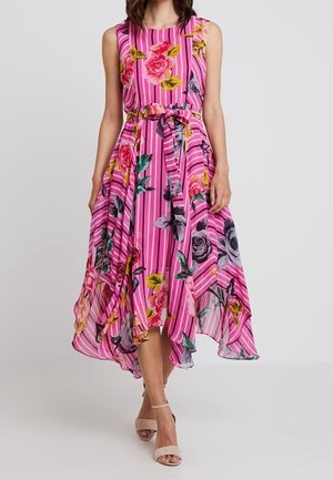 Robe rose sans manches avec des rayures verticales et un grand imprimé floral, taille ceinturée et ourlet asymétrique, portée avec des talons nus à brides.