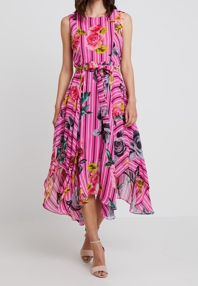 Robe rose sans manches avec des rayures verticales et un grand imprimé floral, taille ceinturée et ourlet asymétrique, portée avec des talons nus à brides.