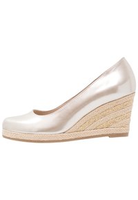 Chaussure compensée beige métallique avec une tige lisse et un intermédiaire en jute tissé. Bout rond et petit talon, avec des accents dorés dans le tissage.