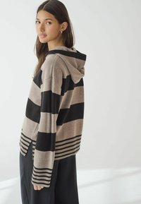 Haut à capuche surdimensionné en beige et noir rayé, présentant un tricot texturé, des poignets côtelés et un ourlet fendu, porté sur un pantalon large noir.
