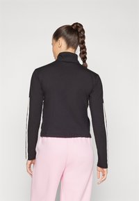 Črn dolg rokav s visokim ovratnikom, ki ima bele stranske črte, gladko teksturo in prilegajočo se zasnovo, v kombinaciji s roza hlačami.