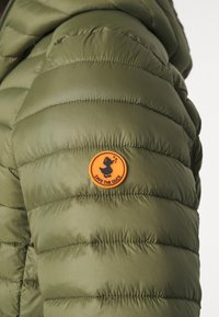 Giacca puffa verde oliva con cuciture orizzontali, dotata di una patch arancione rotonda con la scritta "SAVE THE DUCK" e un logo di anatra.