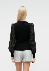 Schwarzer Pullover mit transparenten schwarzen Puffärmeln mit Blumenmuster. Gerippte Bündchen und Saum, getragen mit hellgrauen Shorts.