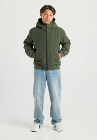 Teen with short hair stands wearing an olive green zip-up hooded jacket, light blue jeans, and white sneakers with black swooshes.