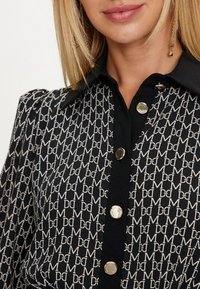 Blusa nera con stampa del logo in beige, con colletto e bottoni argentati lungo la parte anteriore. Tessuto testurizzato con maniche a sbuffo.
