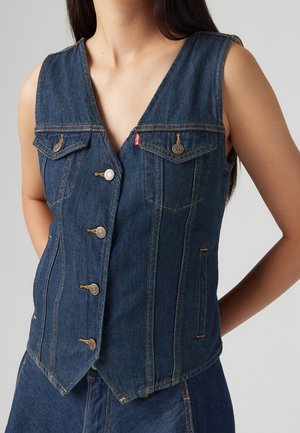 Vrouw draagt een mouwloos blauw denim vest met metalen knopen en voorzakken, weergegeven van nek tot heupen tegen een effen achtergrond.
