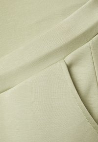 Tecido verde-claro com uma textura suave, apresentando uma seção superior com nervuras e um bolso lateral, exibindo um design limpo e costura simples.