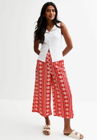 Weiße ärmellose Bluse mit Knöpfen vorne, kombiniert mit roten, gemusterten, weiten Hosen, die weiße Blumenmuster aufweisen. Tragen Sie helle Sandalen dazu.