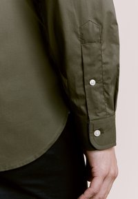 Koszula oliwkowo zielona na guziki, z długimi rękawami, zaokrąglonym dołem, dwoma guzikami na mankiecie oraz gładką fakturą tkaniny.