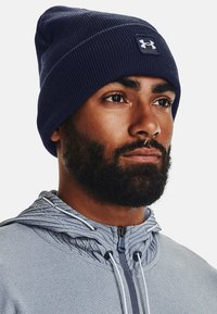 Bonnet en tricot côtelé bleu marine avec un écusson logo blanc, porté avec une veste zippée gris clair en tissu texturé et cordons de serrage ajustables.