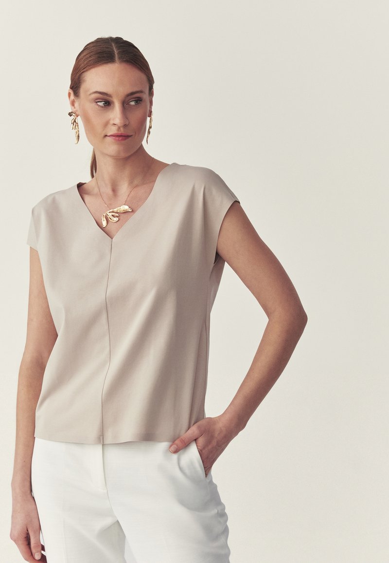 Femme aux cheveux attachés portant un haut beige à manches courtes et encolure en V, un pantalon blanc, des boucles d'oreilles en forme de feuille dorée et un collier assorti, la main dans la poche.