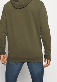 Felpa in cotone verde oliva con tasca a canguro, polsini e orlo a coste. Indossata sopra jeans di denim blu, vista da dietro.