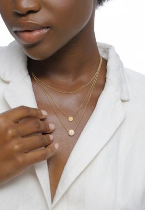 Gros plan d'une personne portant plusieurs colliers en or avec de petits pendentifs ronds, vêtue d'un haut blanc à col, la main près de la poitrine.