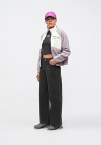 Jeune femme portant une veste polaire violette et blanche, un crop top noir, un pantalon noir à jambes larges, des baskets grises et une casquette violette, debout et regardant sur le côté.