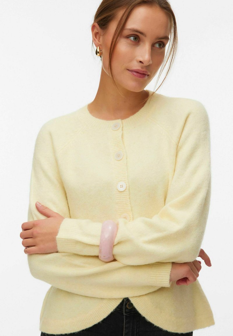 Femme portant un cardigan boutonné jaune pâle, des boucles d'oreilles anneaux dorés et un grand bracelet rose, bras croisés, regardant légèrement sur le côté.