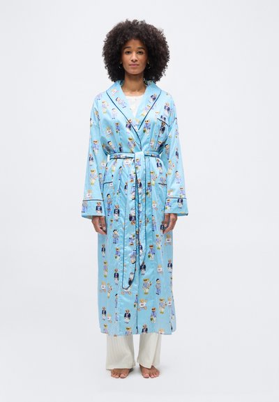 Frau mit lockigem Haar, die einen langen, blau gestreiften Bademantel mit Teddybärmotiven trägt, der in der Taille gebunden ist, barfuß vor weißem Hintergrund steht.