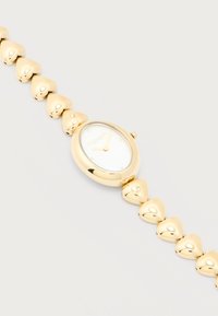 Γυναικείο ρολόι χειρός σε χρυσό τόνο με οβάλ καντράν και κοσμήματα σε σχήμα καρδιάς στη στεφάνη, μινιμαλιστικό καντράν χωρίς αριθμούς, επωνυμία Coach.