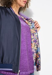 Marinblå bombarjacka med blommönstrad foder, bärs över en texturerad lila topp. Har en silverfärgad dragkedja och ribbade ärmslut.