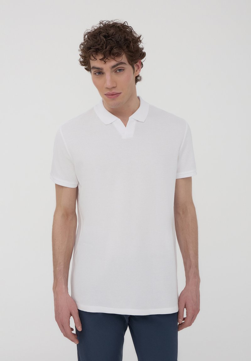Polo bianco a maniche corte, in tessuto texture e con colletto classico. Il design è semplice, senza motivi o dettagli visibili.